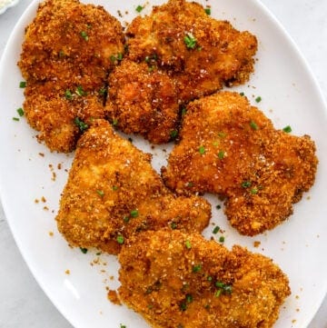 White platter filled with crispy coated chicken thighs with some chopped chives and parmesan cheese in small bowls behind it.