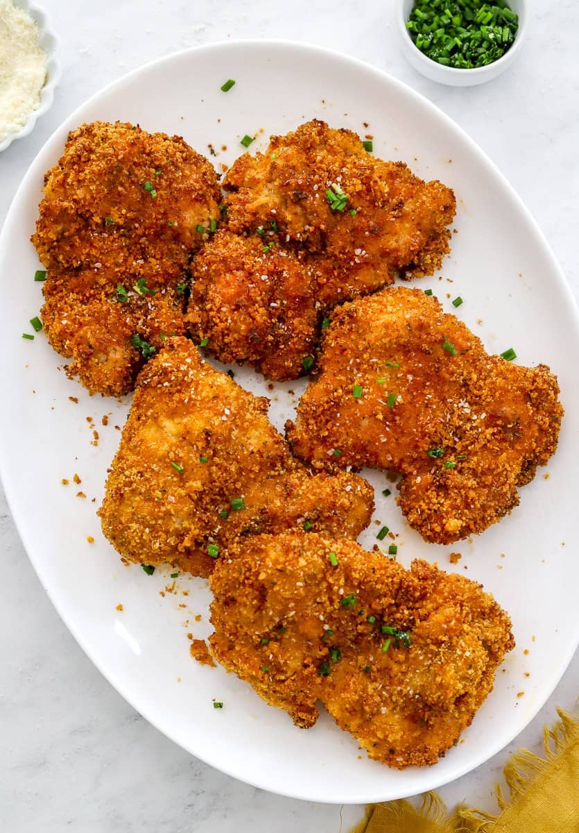 White platter filled with crispy coated chicken thighs with some chopped chives and parmesan cheese in small bowls behind it.