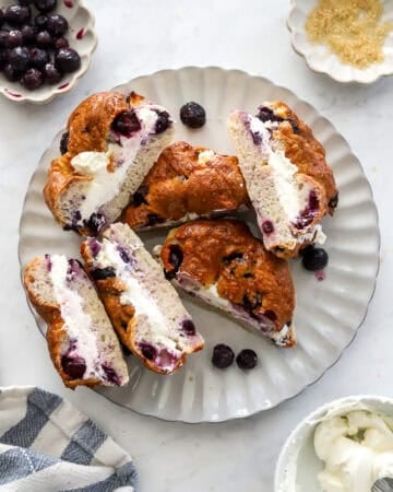 Plate filled with blueberry bagels cream cheese sandwich halves with a bowl of cream cheese in front of it.