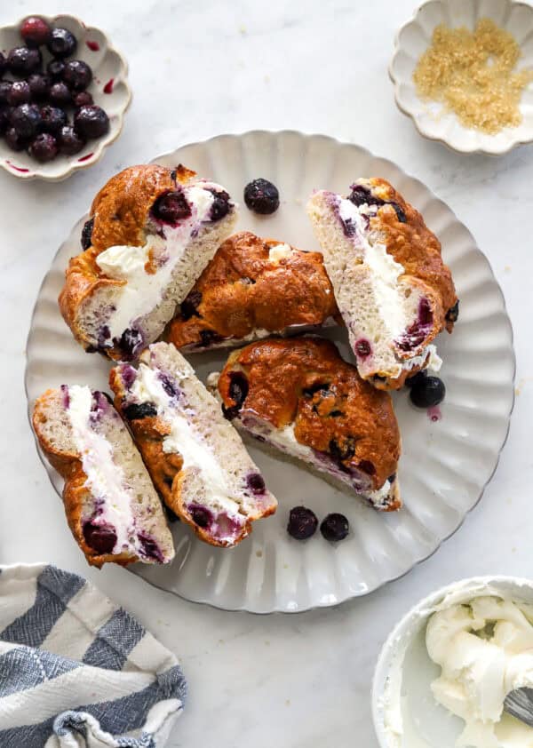 Plate filled with blueberry bagels cream cheese sandwich halves with a bowl of cream cheese in front of it.