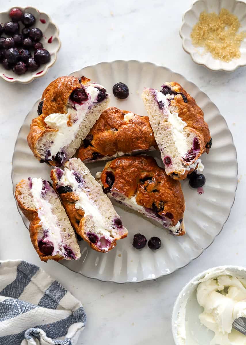 Plate filled with blueberry bagels cream cheese sandwich halves with a bowl of cream cheese in front of it.