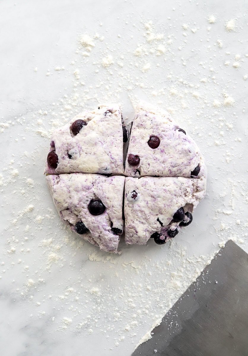 Portioned blueberry bagel dough on a floured surface with a metal cutter in front of it.