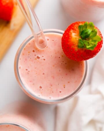 strawberry smoothie in a glass with a clear straw in the glass and a whole strawberry on the glass with more glasses filled with the smoothie mixture next to it.
