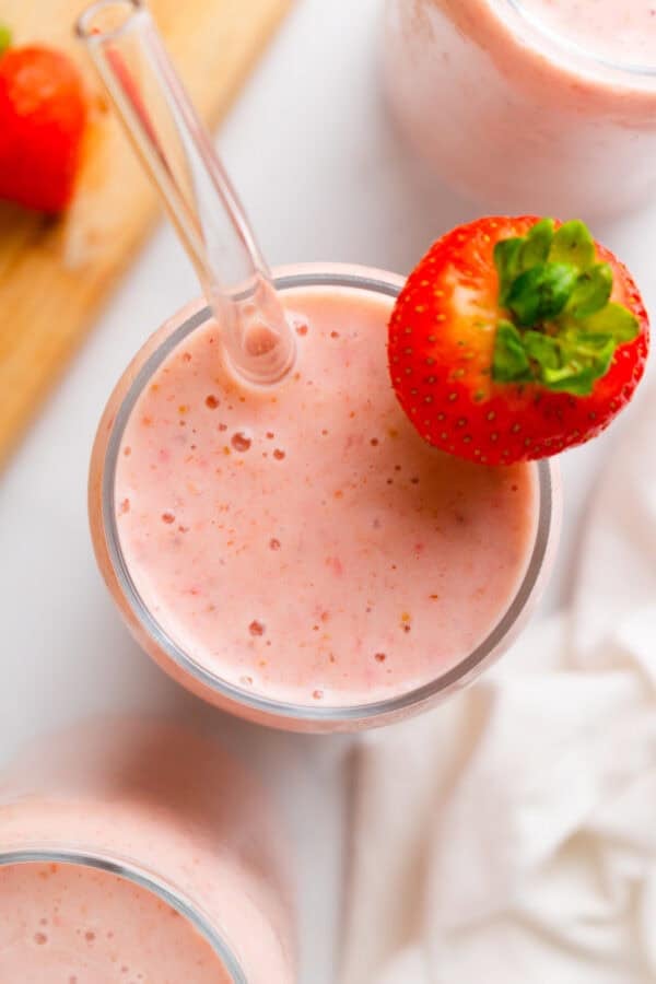 strawberry smoothie in a glass with a clear straw in the glass and a whole strawberry on the glass with more glasses filled with the smoothie mixture next to it.