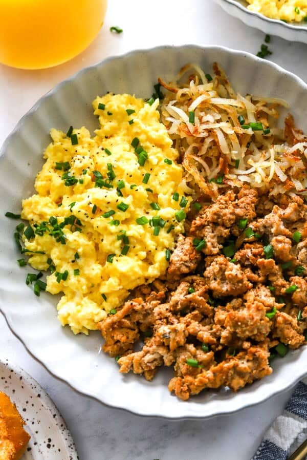 Round bowl filled with cottage cheese eggs, cooked turkey meat and shredded golden potatoes with a glass or orange juice behind it.