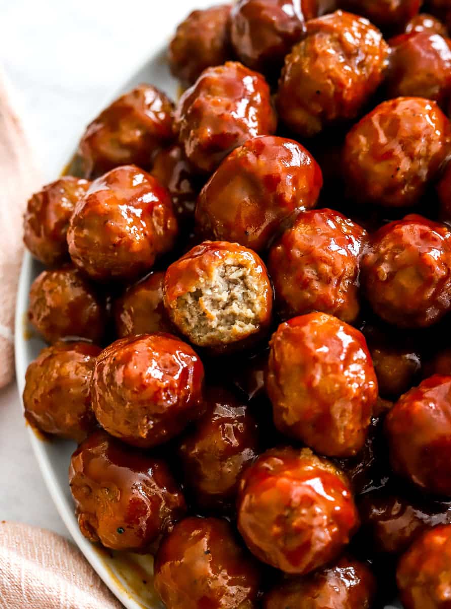 Pile of bbq covered meatballs made in the slow cooker with a bite taken out of one of the meatballs on a plate with a pink towel next to it. 