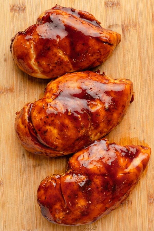 3 cooked chicken breasts covered in bbq sauce on a wood cutting board.
