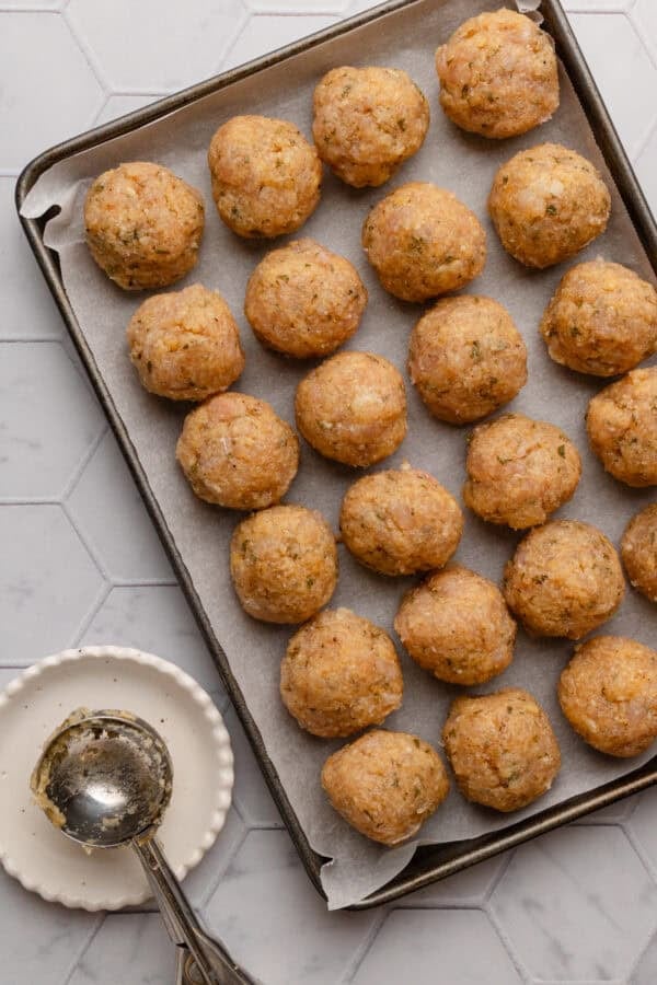Formed uncooked chicken meatballs on a lined pan with a scoop on a plate in front of it.