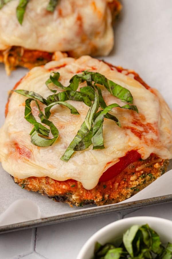 Baked coated mushroom parmesan topped with melted cheese and chopped basil on a lined pan with more chopped basil in a bowl in front of it.