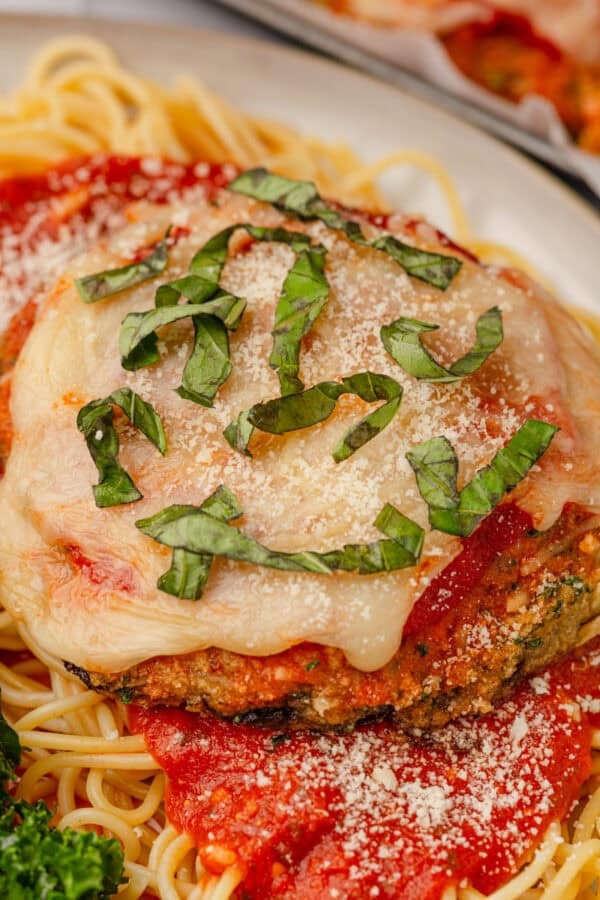 Portobello mushroom parnesan on a plate of spaghetti with red sauce, topped with chopped basil.