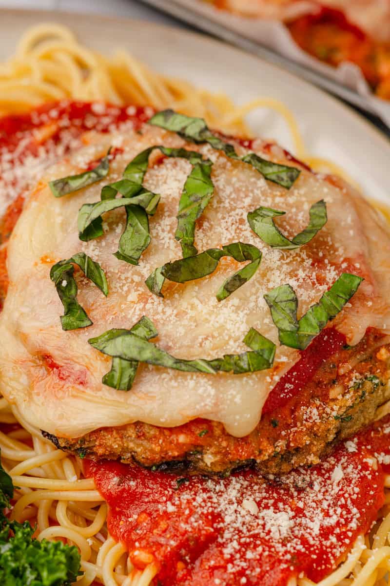 Portobello mushroom parnesan on a plate of spaghetti with red sauce, topped with chopped basil.