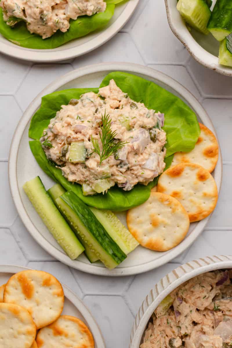 Round plate with a lettuce cup filled with creamy salmon salad with crackers and cucumber on the plate with it and more behind it. 