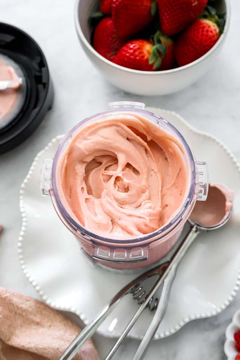 Strawberry blended ice cream in Ninja Creami container on a plate with ice cream scoop next to it.