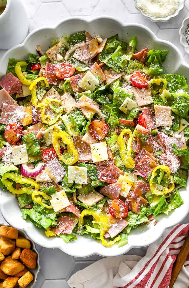 Large salad bowl filled with Italian grinder salad topped with grated parmesan cheese and Italian grinder dressing with a small bowl of parmesan cheese behind it. 