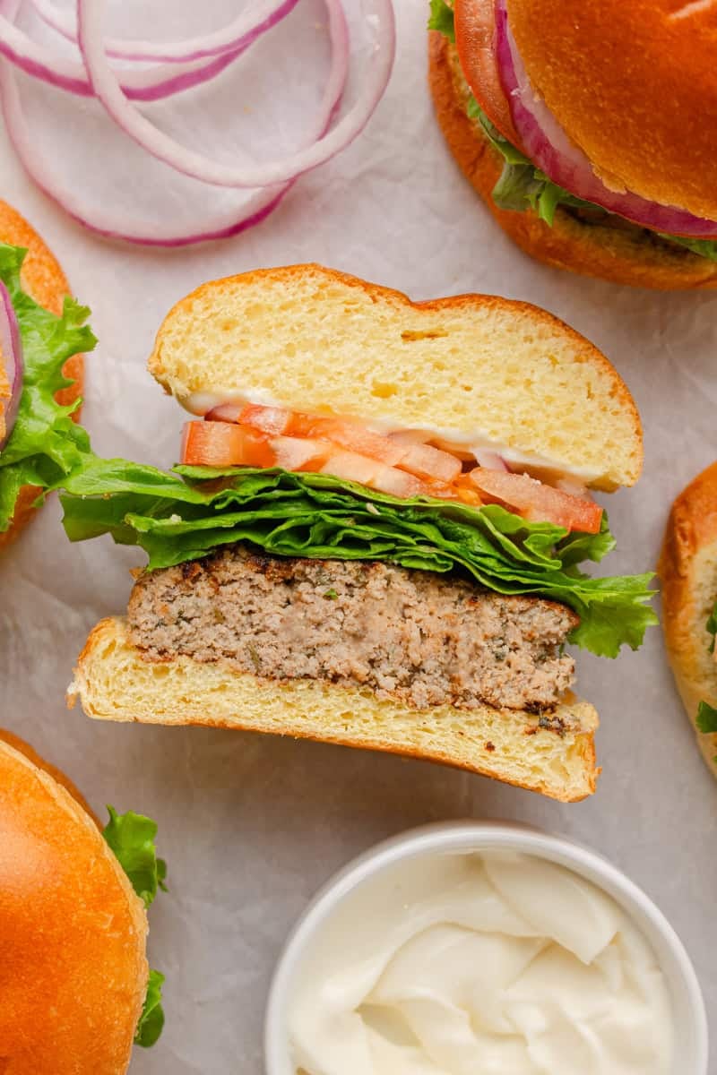 Cut turkey burger on a roll with lettuce and tomato with more burgers around it and a bowl of mayo in front of it. 