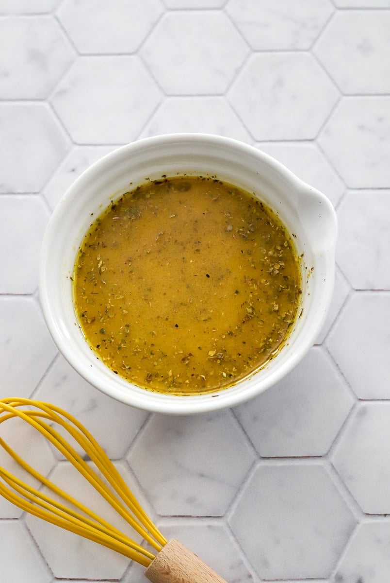 Grinder salad dressing in a white bowl with a yellow whisk in front of it. 