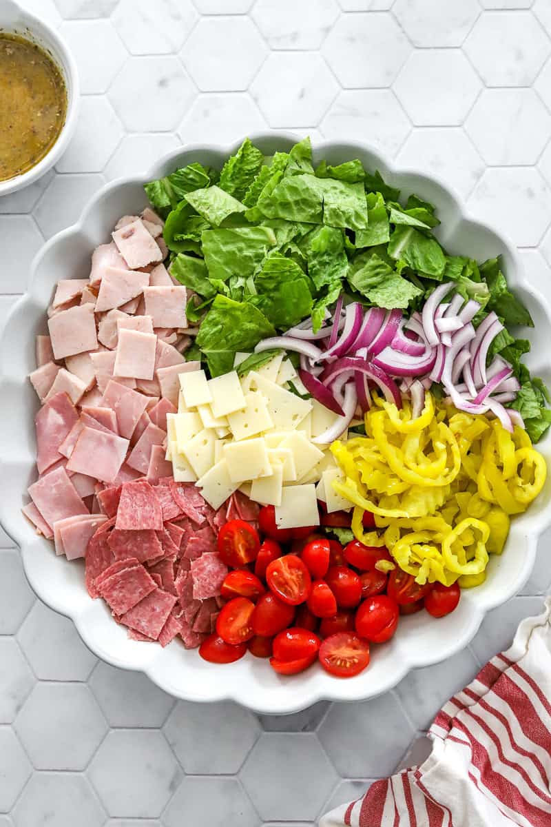 Large salad bowl filled with chopped lettuce, sliced cherry tomatoes, onion, banana peppers, chopped cheese and deli meat in it. 