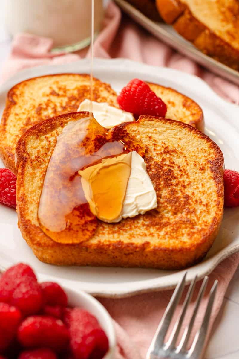 Protein French toast on a plate topped with butter and syrup with raspberries around it. 