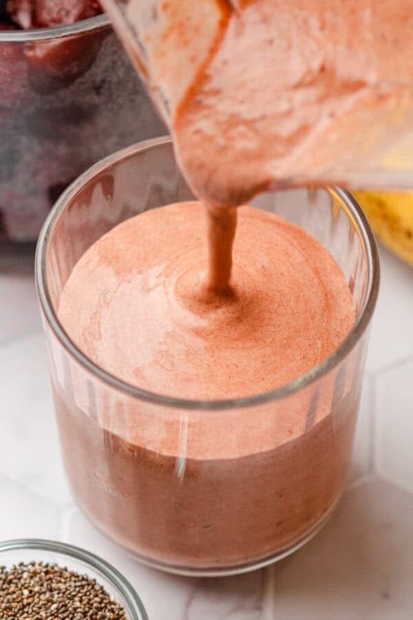 Pitcher pouring a pink smoothie with cherries into a glass with a bowl of chis seeds in front of it.