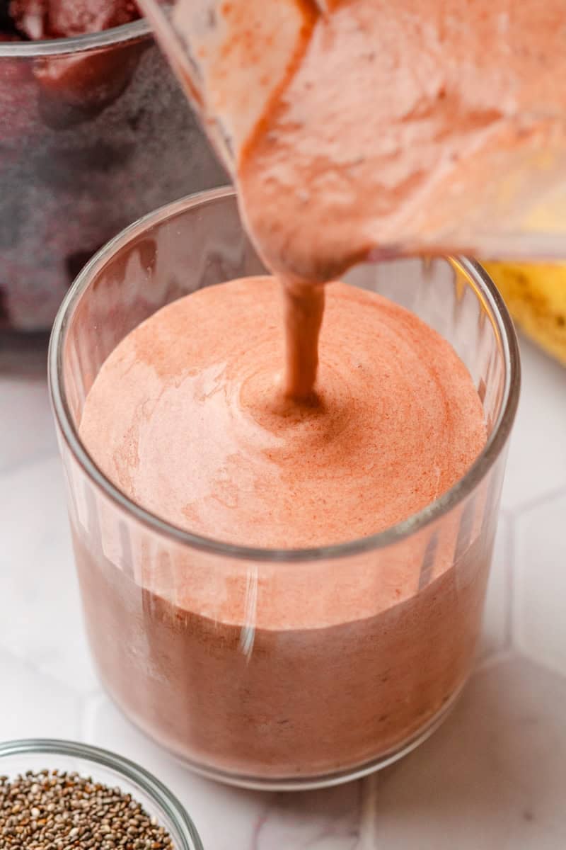 Pitcher pouring a pink smoothie with cherries into a glass with a bowl of chis seeds in front of it.