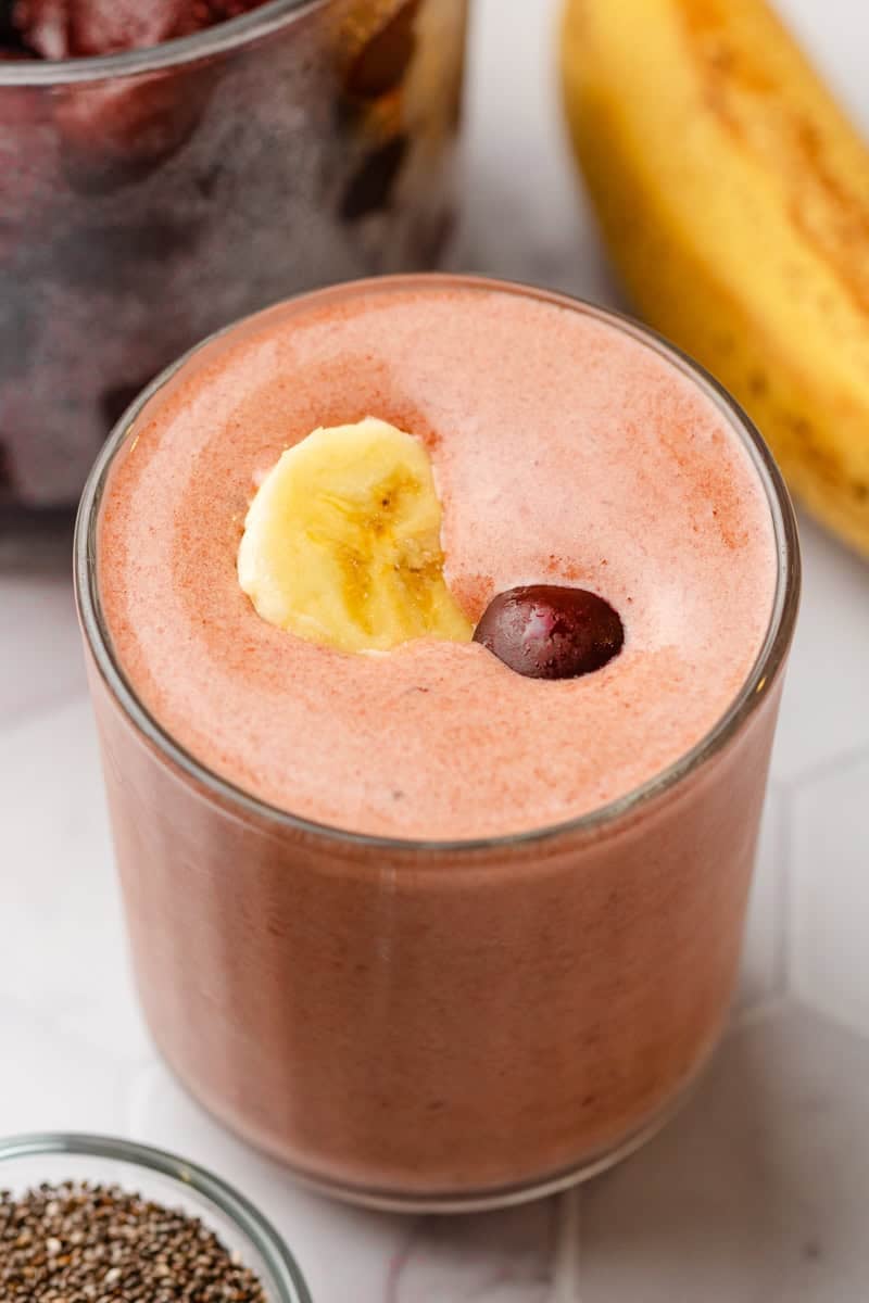 Glass filled with a pink cherry blended drink with a bowl of cherries and a banana behind it.