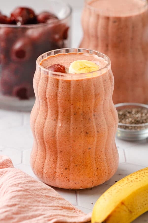 Two smoothies in glasses with a banana in front of them and a bowl of cherries behind them.