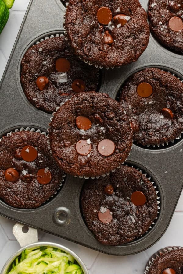 Baked chocolate muffins with zucchini in a muffin pan with one of the muffins on top of the pan.