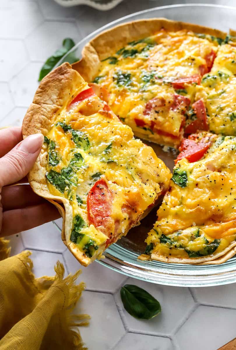 Hand pulling a slice of quiche out of the pan with more baked tortillas quiche behind it. 
