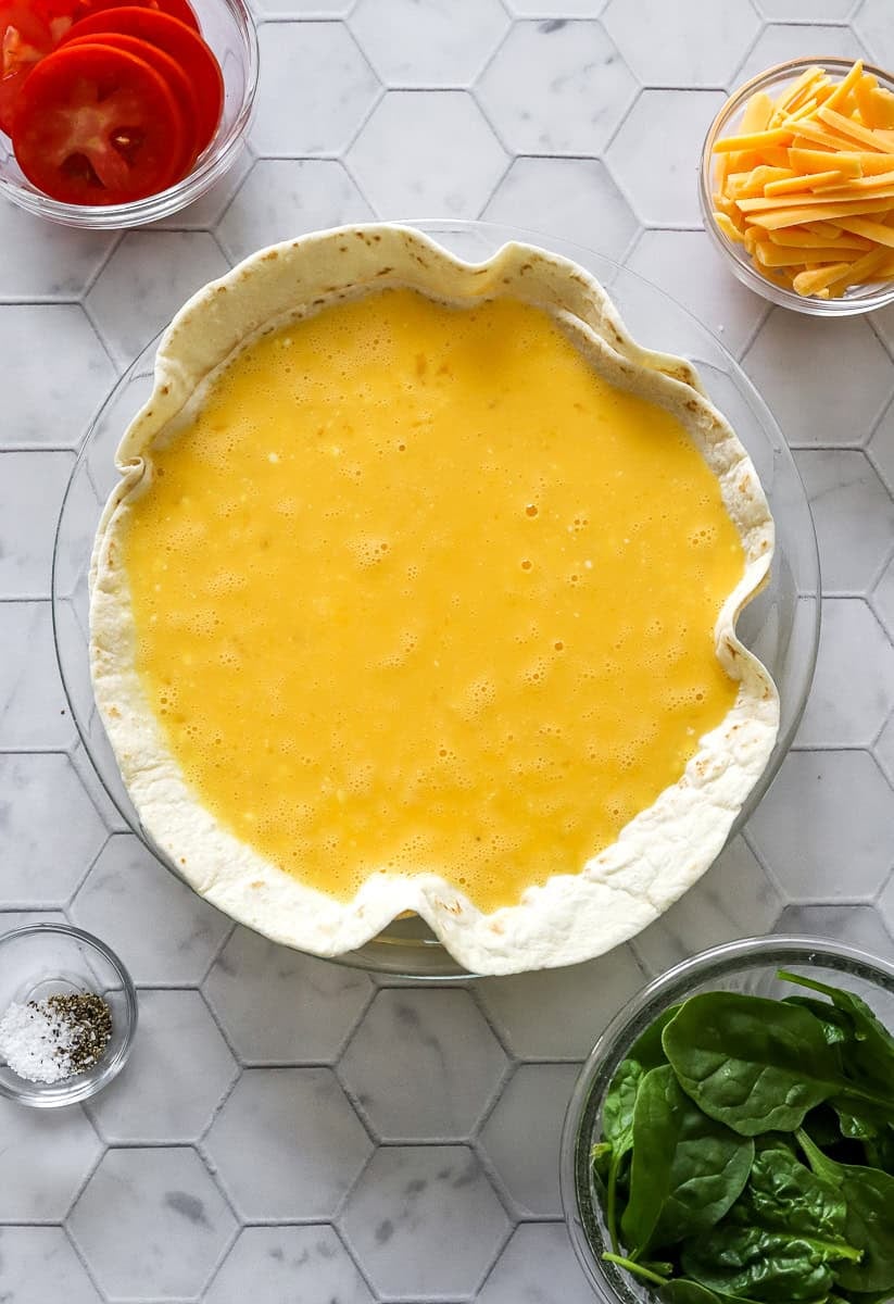 Egg filling in tortilla in a pie dish with a bowl of spinach in front of it and a bowl of sliced tomatoes behind it.