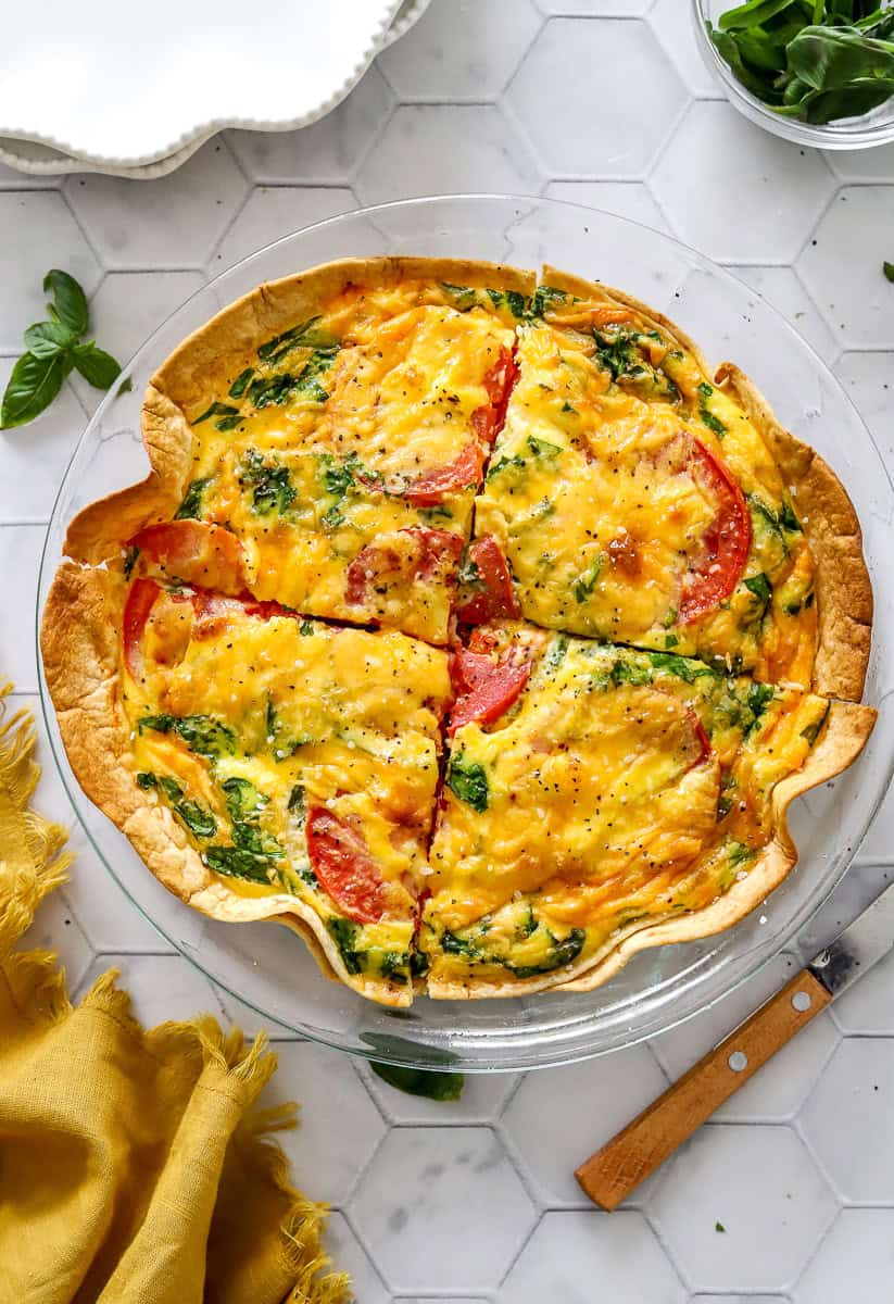 Baked tortilla quiche with spinach and tomatoes cut into portions in a glass pie dish with a knife and yellow dish towel in front of it.