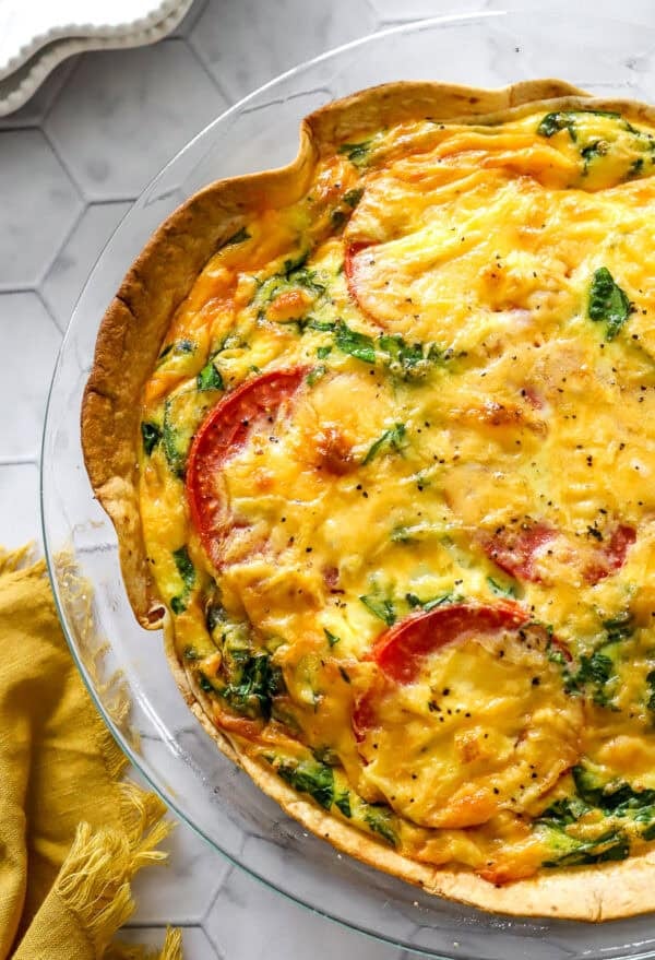 Baked quiche in a tortilla crust with spinach, tomatoes and cheese with a yellow dish towel next to it.