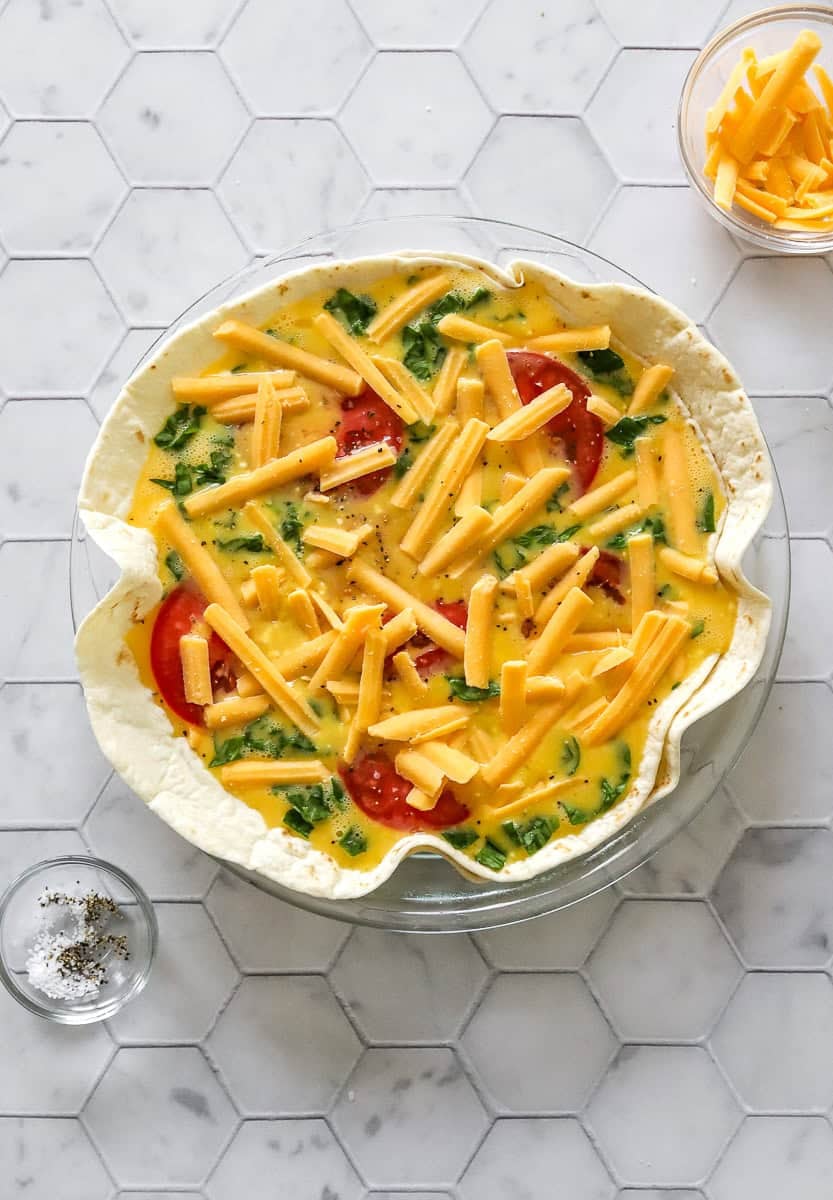 Large tortillas arranged in a glass pie dish with whisked egg and veggie filling in it topped with shredded cheddar cheese.