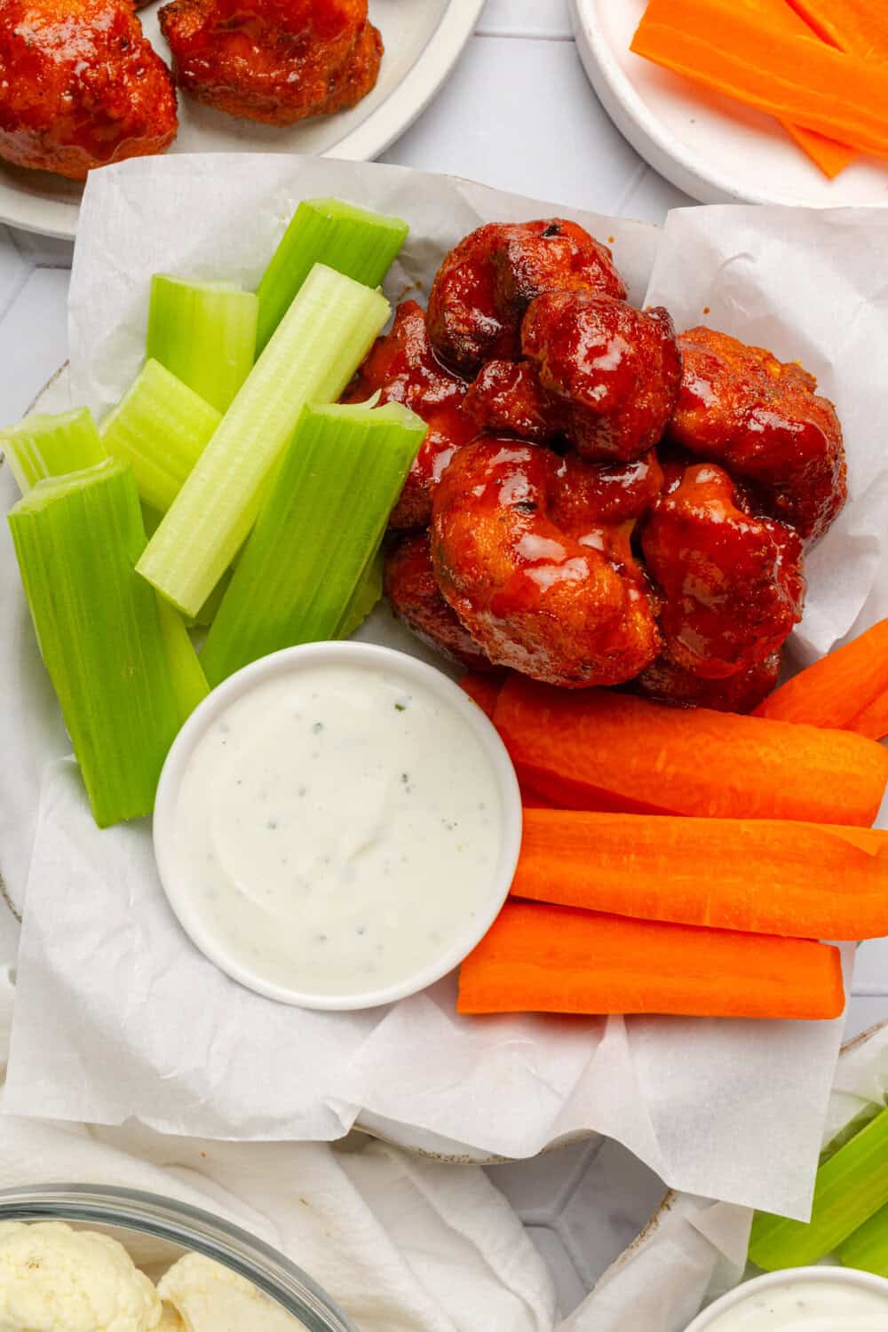 Plate filled with hot an d spicy coated cauliflower next to a dish of creamy dip and cut celery and carrot sticks with more around it.