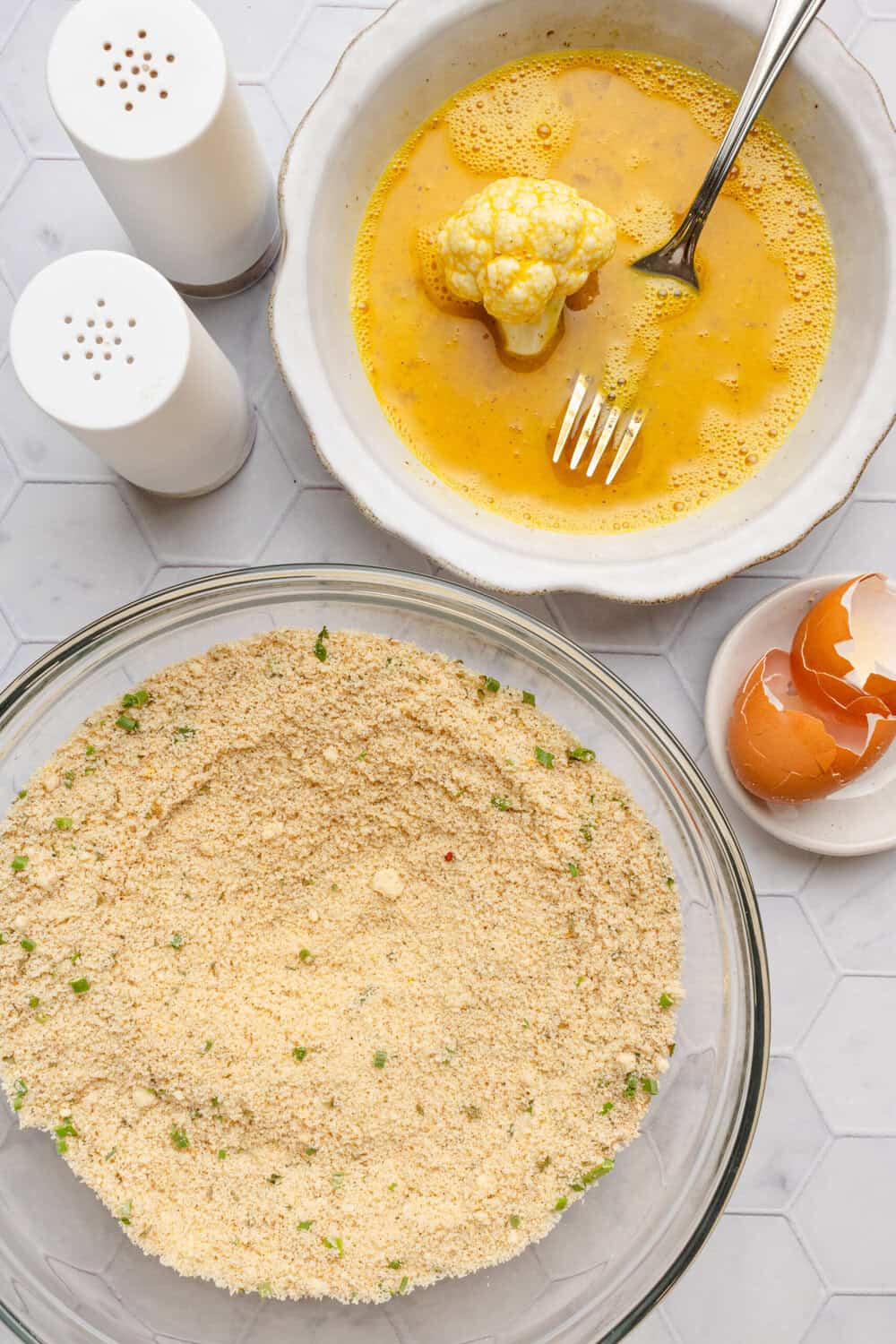 Shallow dish of breading with another shallow dish with whisked egg and a cauliflower floret in it.