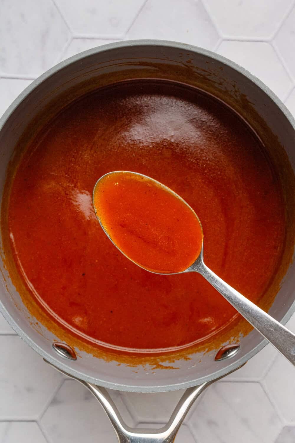 Homemade buffalo sauce in a pot with a spoon lifting some of the sauce out of the pot.