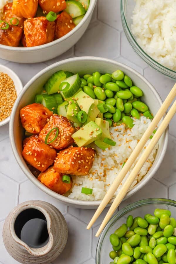 Air fryer teriyaki salmon bites over rice in a bowl with veggies and chopsticks on the bowl with more ingredients around it.