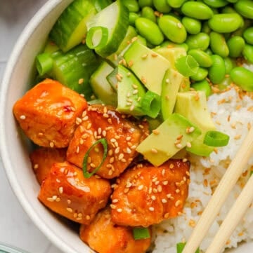 Air fryer teriyaki salmon bites over rice in a bowl with veggies and chopsticks on the bowl with more ingredients around it.