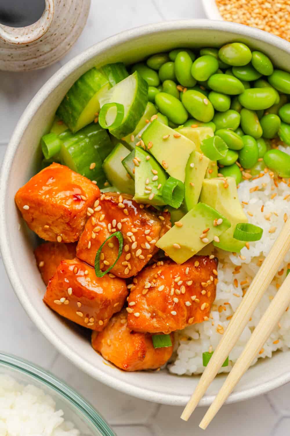 Air fryer teriyaki salmon bites over rice in a bowl with veggies and chopsticks on the bowl with more ingredients around it.