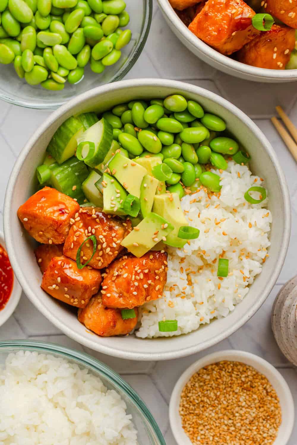 Rice salmon bowl with edamame and avocado with more ingredients around it. 