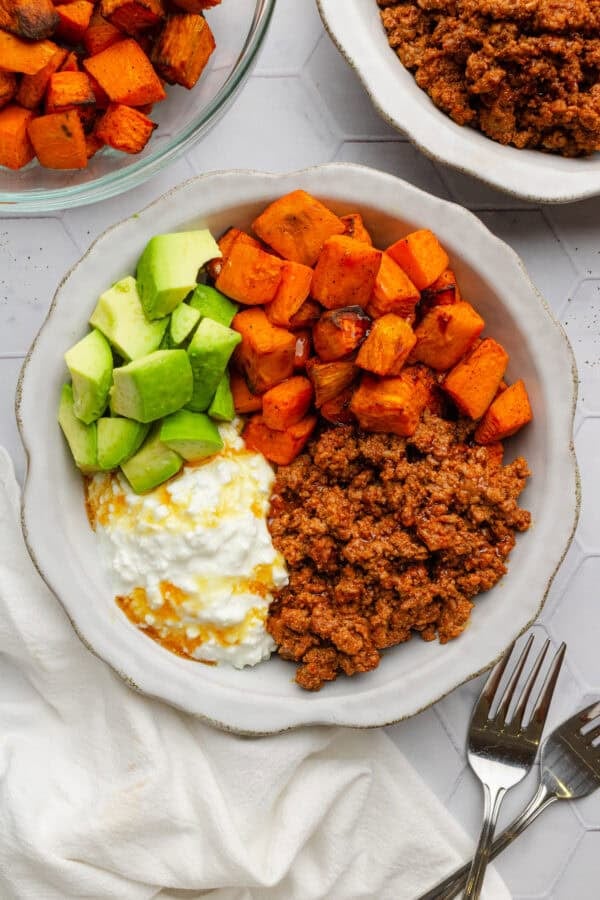 Bowl filled with cooked beef, air fried sweet potato cubes, chopped avocado and cottage cheese topped with hot honey with more ingredients behind it.