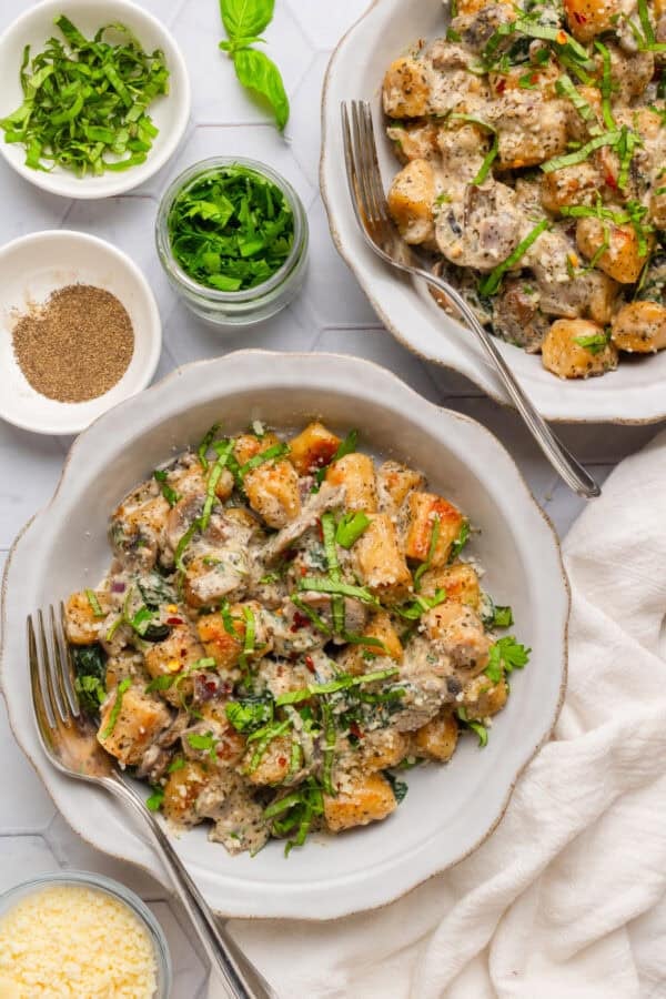 Two bowl of cooked creamy spinach and mushroom gnocchi with more ingredients next to it.