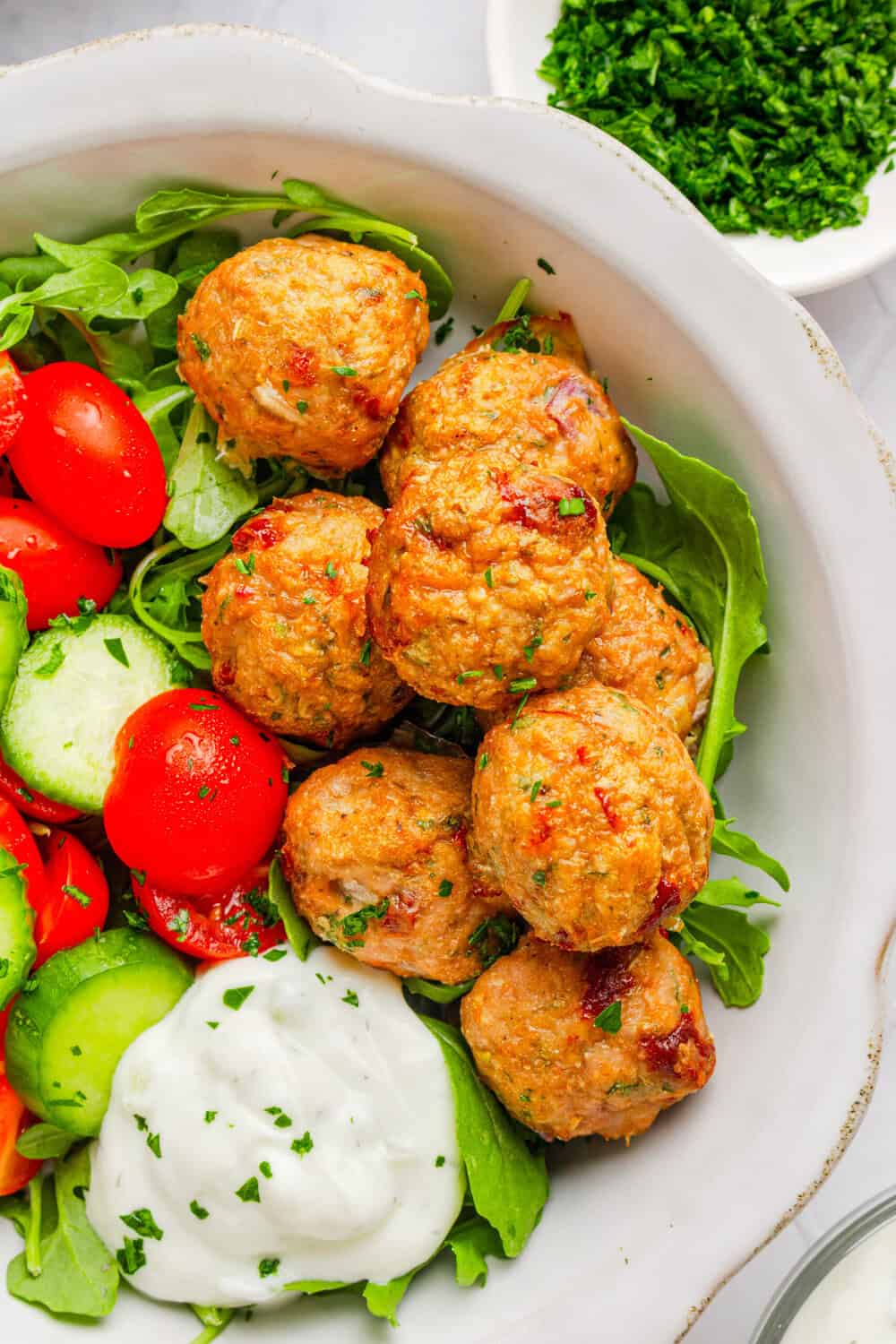 Healthy greek meatballs in a white bowl on top of chopped cucumbers and tomatoes with a creamy white sauce next to them. With a slice of lemon and a few pieces of herbs next to the bowl. 