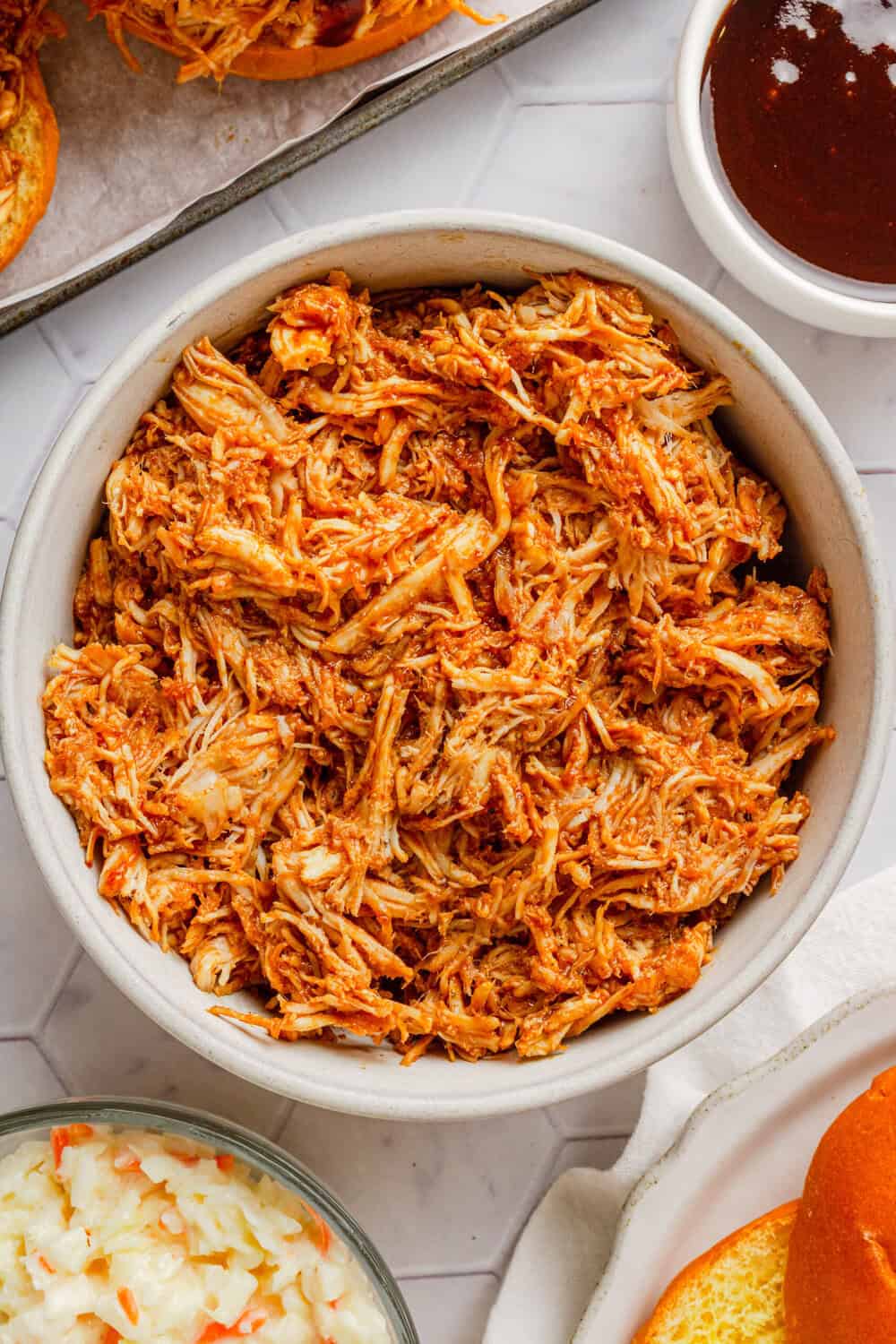 Shredded cooked BBQ coated chicken breast in a bowl with more ingredients around it. 