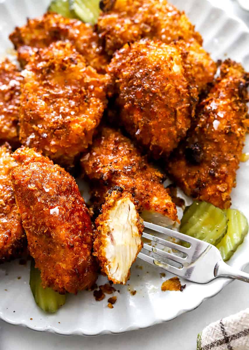 Plate of air fried hot honey chicken with a piece cut on a fork to see the inside, with pickle on the plate with it. 