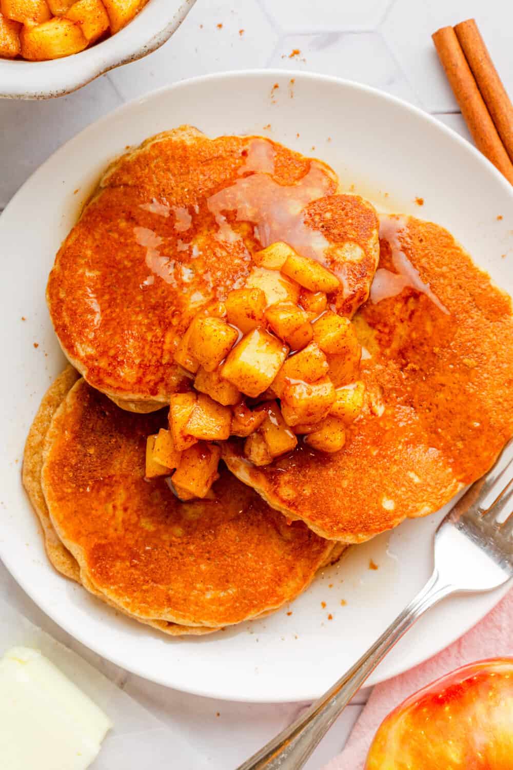 White plate of cooked cinnamon pancakes with chopped cooked apples and syrup on top of the pancakes with more cooked apples behind it.