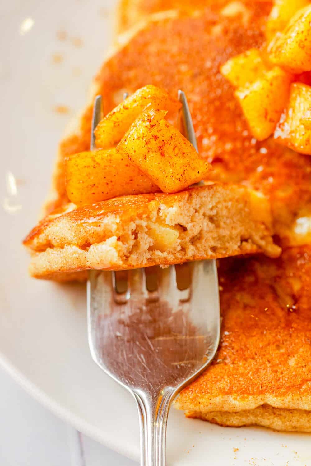 Piece on apple pancakes on a fork with apple pieces on the fork with it and more pancakes behind it. 