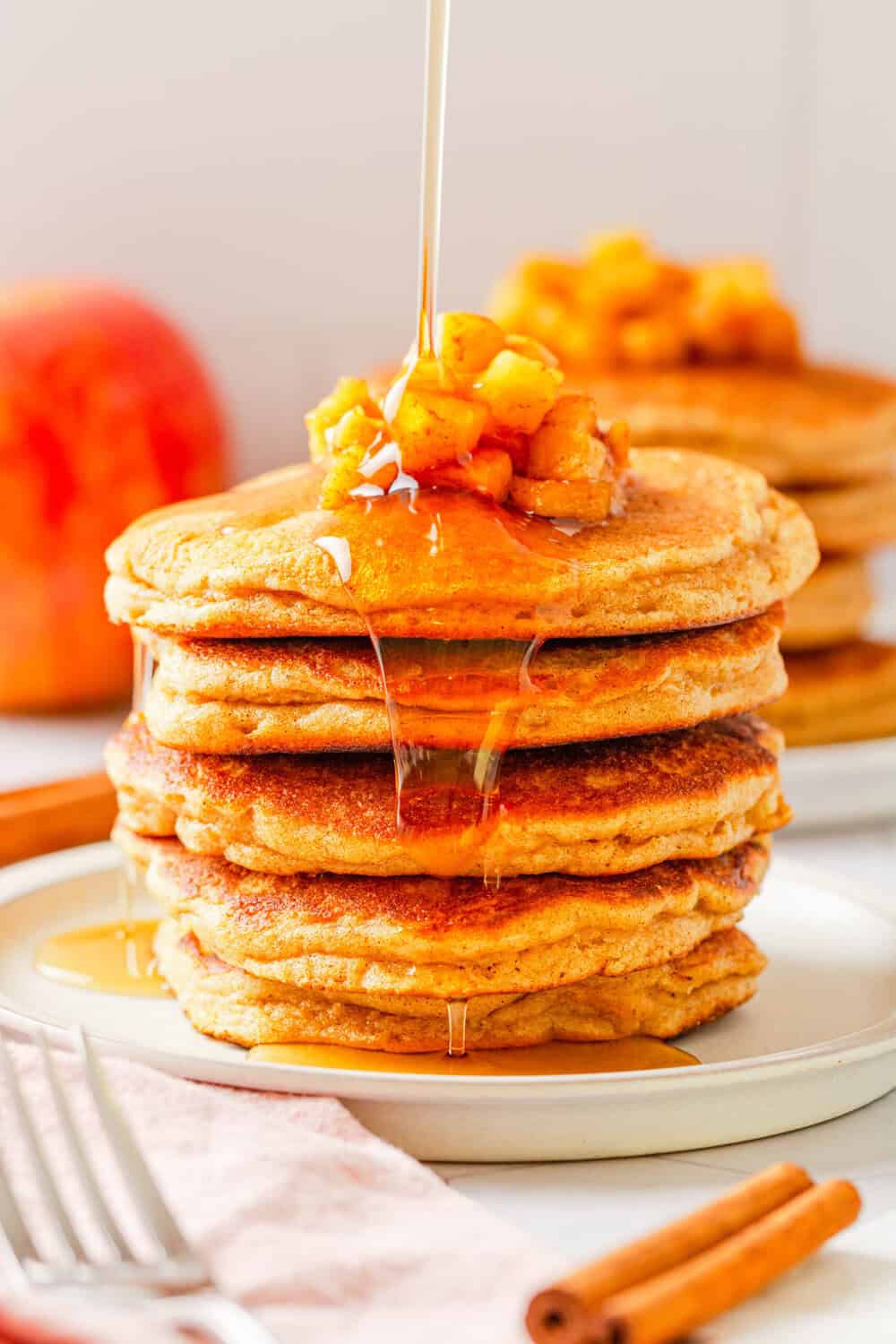 Stack of gluten free apple pancakes with chopped cooked apples on top and maple syrup drizzled with more pancakes behind it. 