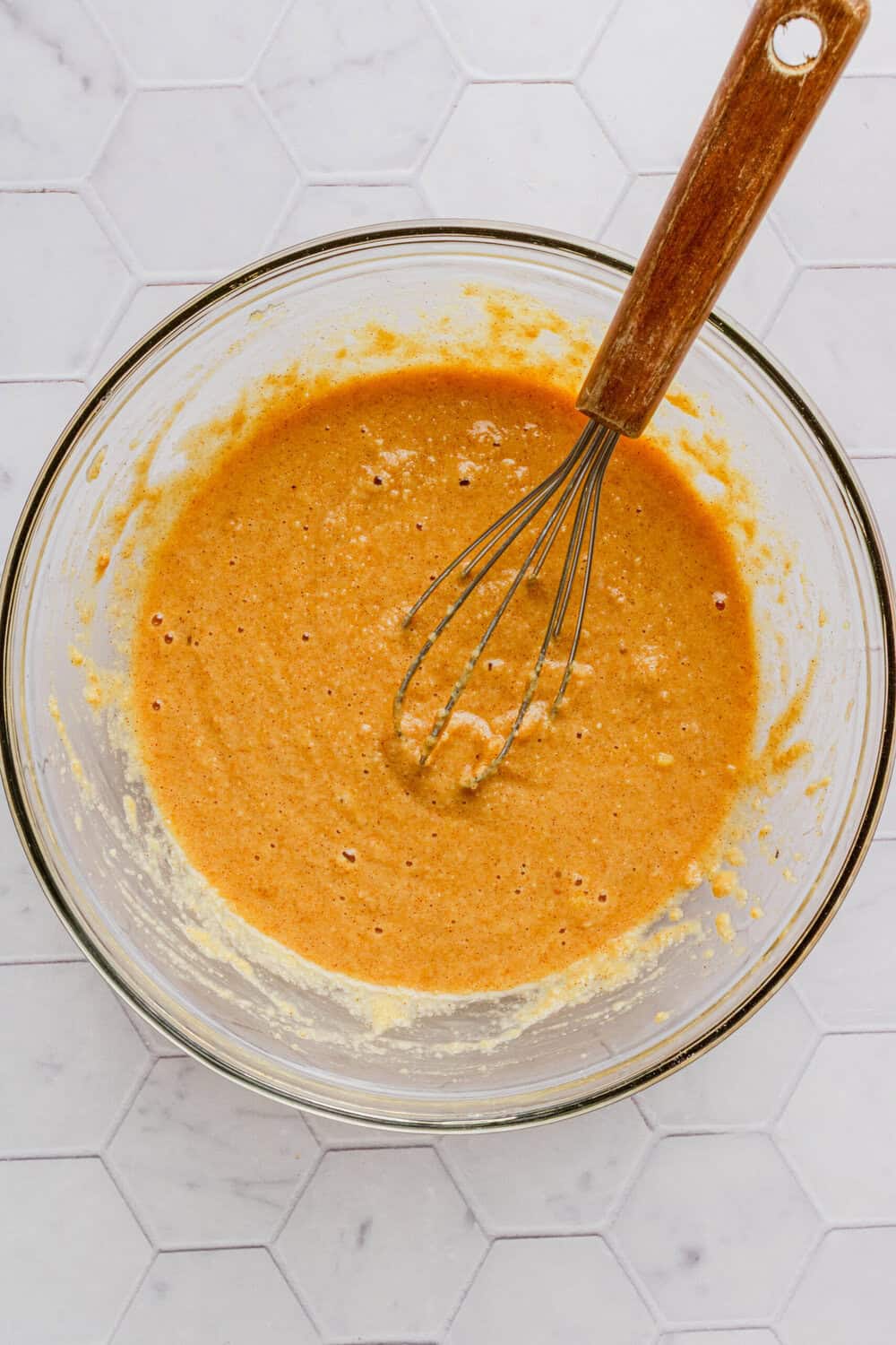 Glass mixing bowl with wet and dray pancakes ingredients blended together in it with a whisk in bowl. 