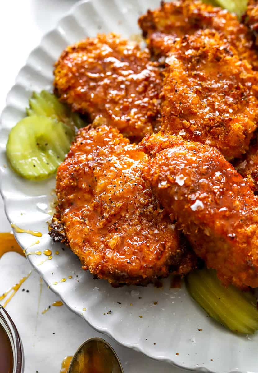 Plate with pieces of cooked crispy coated chicken drizzled with hot honey with pickles on the plate with it. 