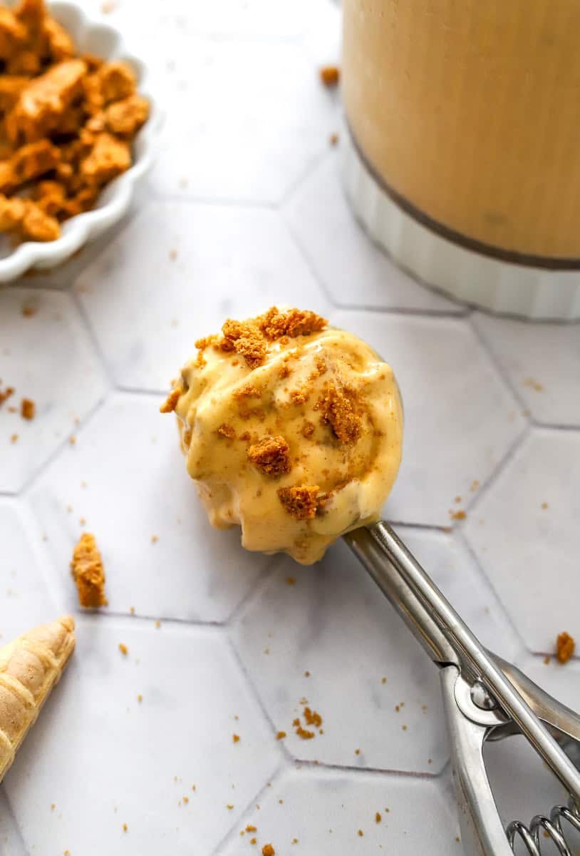 Ice cream scoop with a scoop of pumpkin Ninja Creami ice cream with crushed cookies in it with more crushed cookies in a bowl behind it.